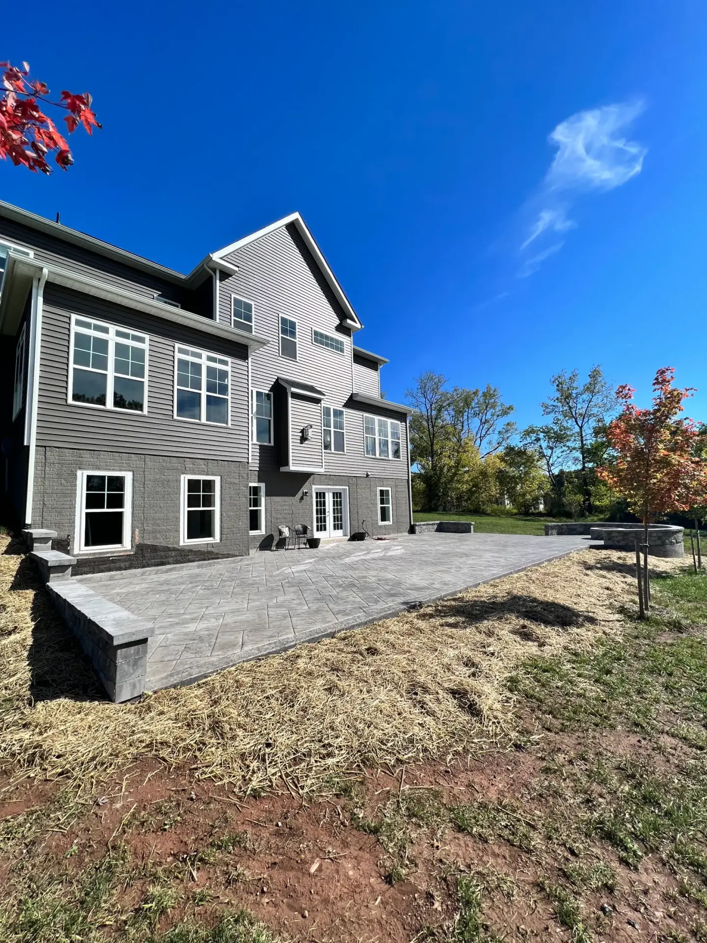 Modern gray, multi-story home with large white-framed windows and a paved backyard patio under a clear blue sky.